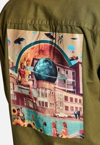 Maglietta verde oliva con una vivace stampa grafica che presenta pianeti, edifici e figure in una piscina, decorata con girasoli ed elementi retro.