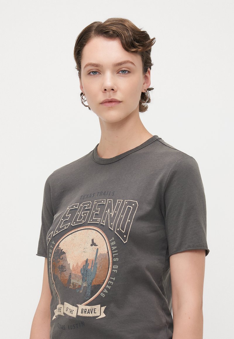 Camiseta de algodón gris con un estampado gráfico que representa una escena del desierto con cactus y texto: "Leyenda, Rutas de Texas," escote redondo, mangas cortas.