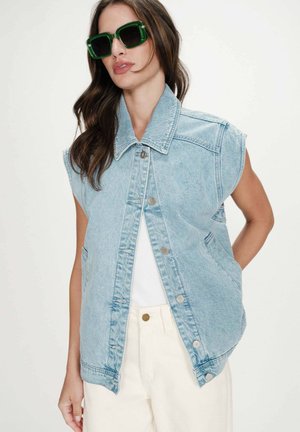 Femme portant un gilet en jean bleu clair sans manches, un haut blanc, un pantalon crème et de grandes lunettes de soleil vertes, aux cheveux bruns lâchés.