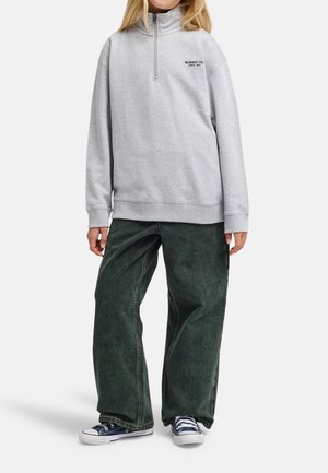 Person trägt hellgraues Sweatshirt mit Viertel-Reißverschluss und Text, lockere dunkelgrüne Hose, marineblaue Turnschuhe, steht vor weißem Hintergrund.