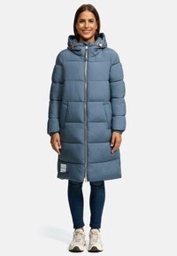 Gewatteerde lange jas in het blauw, met een ritsluiting en capuchon. Zacht van textuur met horizontale stiksels en een logopatch aan de onderkant. Draagt over jeans.