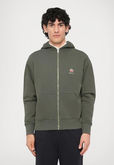 Homme aux cheveux bouclés portant un hoodie zippé vert olive avec des poches avant et un petit logo sur la poitrine, associé à un pantalon noir.
