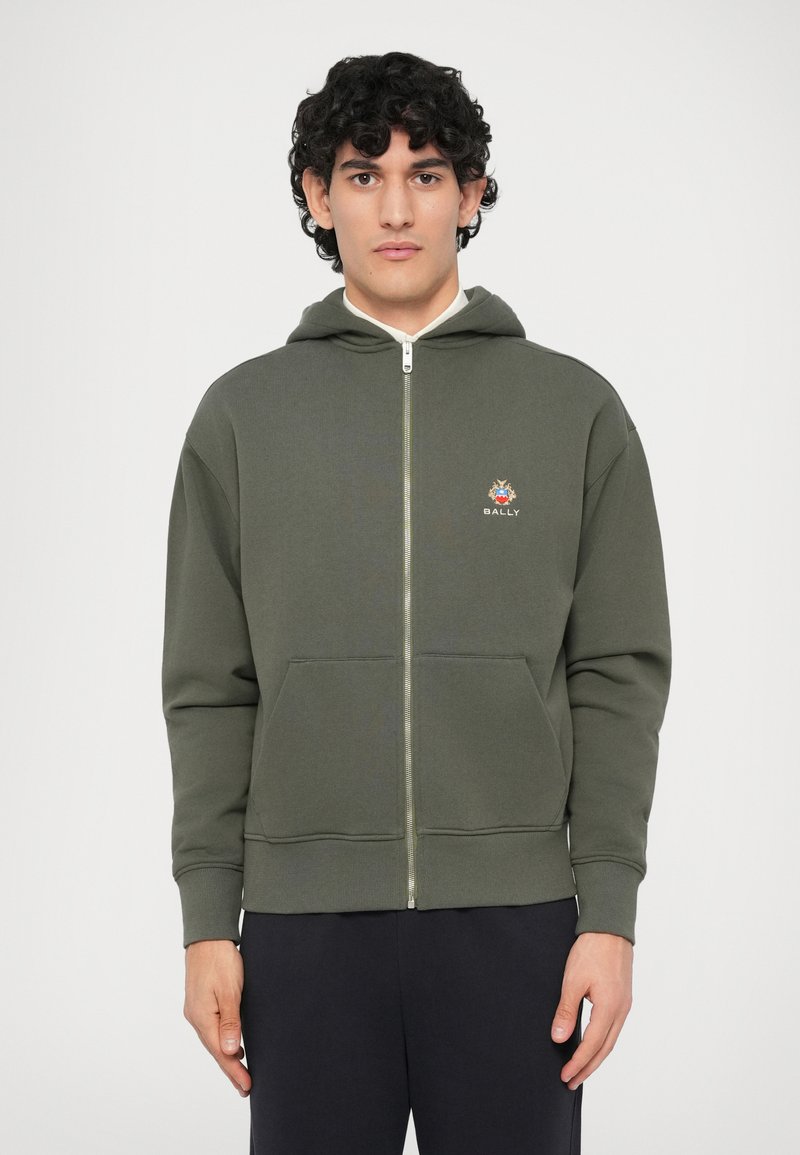 Hombre con cabello rizado que lleva puesta una sudadera con cremallera en verde oliva, con bolsillos frontales y un pequeño logo en el pecho, combinada con pantalones negros.
