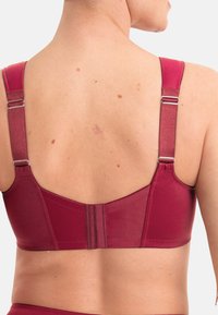 Soutien-gorge en tissu rouge avec bretelles réglables, panneau arrière texturé et fermeture à crochets. Matériau lisse avec un léger éclat et des coutures minimales.