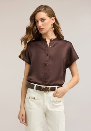 Frau trägt eine kurzärmelige dunkelbraune Satinbluse, die in weiße hoch taillierte Hosen mit einem goldenen Schnallen-Gürtel und dekorativen Vorder taschen gesteckt ist.