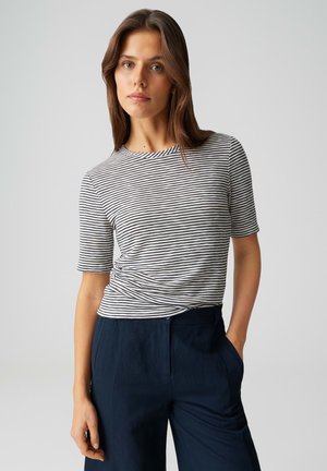 Junge Frau mit braunen Haaren, die ein schwarz-weiß gestreiftes kurzärmeliges Shirt und dunkelblaue Hochbundhosen trägt, mit einer Hand in der Tasche stehend.