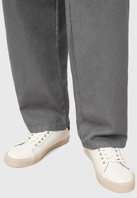 Baskets en toile blanches avec une finition texturée, semelles en caoutchouc plates et œillets en argent, portées avec un pantalon gris ample.