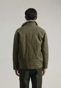 Chaqueta verde oliva con cuello de borrego, diseño estructurado, costuras visibles y detalles de botones. Se lleva con pantalones verdes a juego.