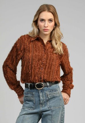 Jeune femme aux longs cheveux blonds porte une chemise texturée couleur rouille rentrée dans un jean bleu taille haute avec de grandes poches avant.