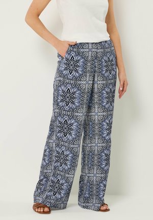 Donna che indossa pantaloni a gamba larga con motivo a piastrelle floreali blu e nere, abbinati a una maglietta bianca e sandali marroni con punta aperta.