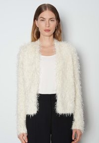 Cardigan bianco e morbido con maniche lunghe, caratterizzato da un materiale testurizzato e peloso. Indossato sopra una maglietta bianca e abbinato a pantaloni neri.