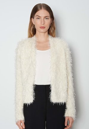 Weißer, flauschiger Cardigan mit langen Ärmeln, aus einem strukturierten, samtigen Material. Über einem weißen Oberteil getragen und mit schwarzen Hosen kombiniert.