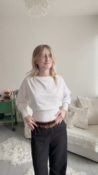 White, loose-fitting sweater with ribbed hem, paired with black high-waisted trousers and a brown belt, standing in a bright indoor setting.