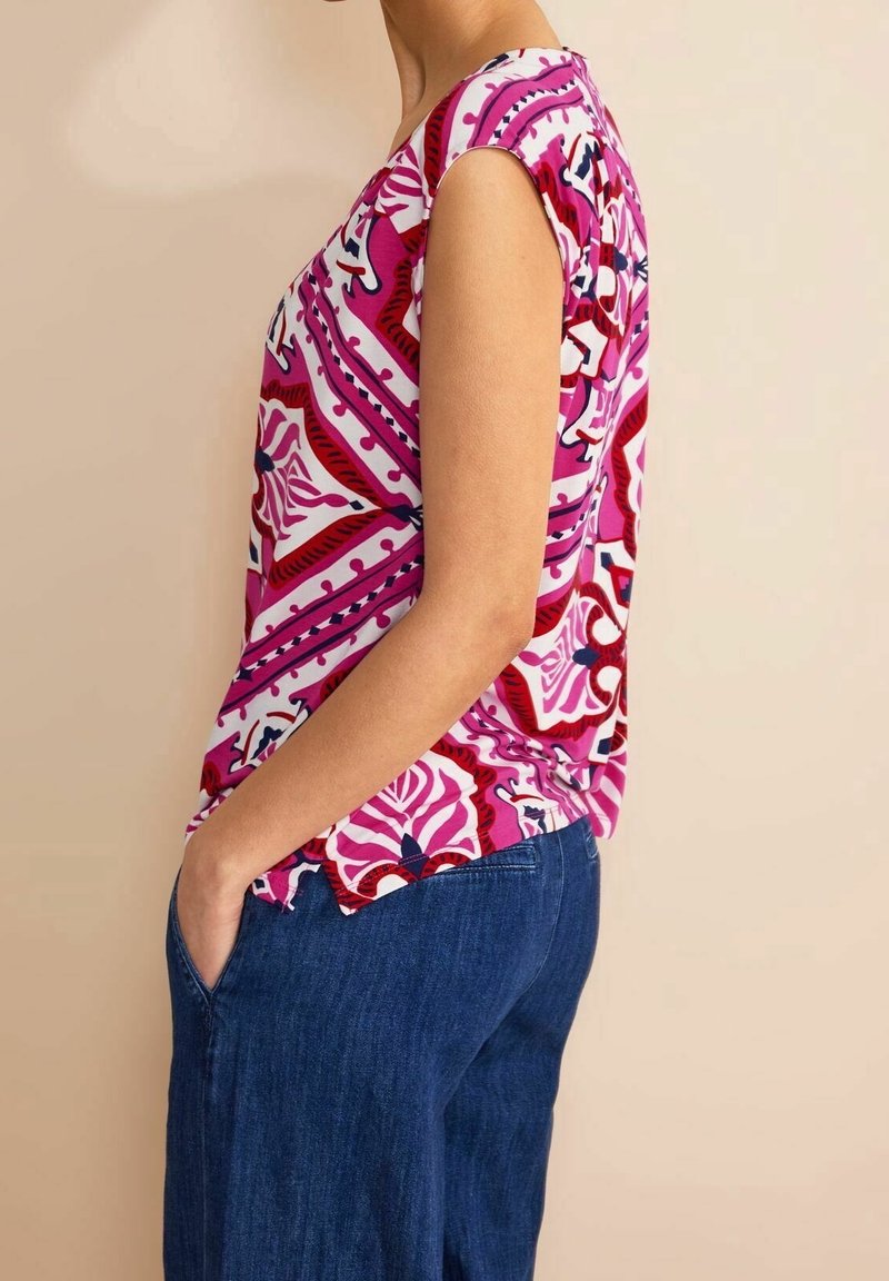 Kortärmad topp i livligt rosa med abstrakta blommönster, tillverkad av ett mjukt tyg. Matchad med blå vida jeans.