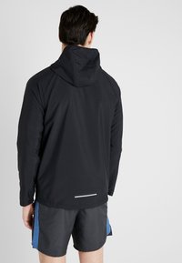 Zwarte jas met een capuchon, gemaakt van lichtgewicht materiaal. Voorzien van raglansmouwen en een reflecterende strook aan de achterkant. Gedragen over een korte broek.