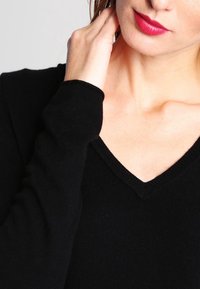 Schwarzer, enganliegender V-Ausschnitt-Pullover mit gerippter Textur und langen Ärmeln. Nahaufnahme, die den Ausschnitt und die Ärmeldetails hervorhebt.
