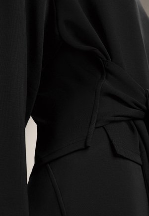 Close-up of black fabric garment featuring a gathered or knotted detail at the waist and visible seam stitching.