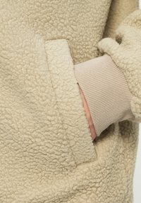 Strato esterno in pile beige con superficie testurizzata, caratterizzato da un polsino lungo fino al polso in un beige chiaro e liscio, e un dettaglio di tasca laterale.