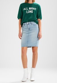 Grön paljettkortärmad sweatshirt med vit text, matchad med en ljusblå denimpennkjol och vita ankelstövlar.