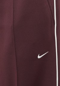 Bordo sportiniai džinsai su baltomis šoninėmis juostelėmis. Priekyje yra mažas baltas Nike logotipas. Lygus, tekstūruotas audinys, vidutinio aukščio dizainas.