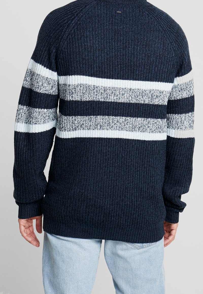 Granatowy sweter z dzianiny z białymi i jasnoszarymi paskami wzdłuż środkowej części. Posiada teksturowany, prążkowany wzór oraz długie rękawy.
