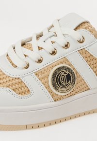 Hvit sneaker med vevde beige detaljer, med gullhull og et rundt logo-plakett. Glatt tekstur med gummisåle.