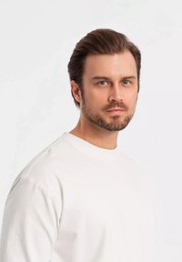 Camisa blanca de manga larga con un escote redondo, fabricada en un tejido suave, de diseño minimalista. El sujeto tiene el cabello castaño claro y barba, con una expresión neutral.