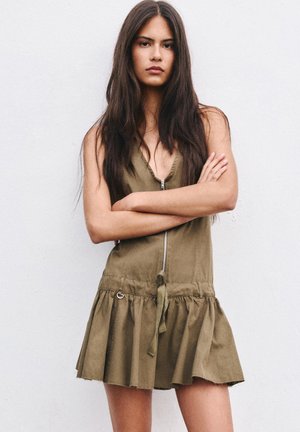 Młoda kobieta z długimi, ciemnymi włosami stoi ze skrzyżowanymi ramionami, ubrana w bezrękawową oliwkowozieloną sukienkę z przednim zamkiem błyskawicznym i falbanką na spódnicy.