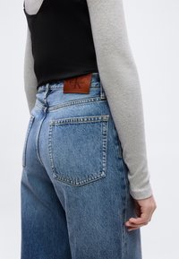 Άτομο που φοράει ανοιχτό μπλε τζιν ψηλής μέσης με καφέ δερμάτινο ταμπελάκι Calvin Klein και μαύρο τοπ επάνω από γκρι μακρυμάνικο πουκάμισο.