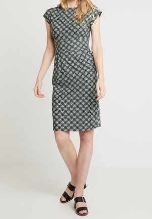 Kobieta w krótkiej, sięgającej kolan sukience w wzory z krótkim rękawem, nosząca czarne sandały z odkrytymi palcami, stojąca na tle jasnego, gładkiego tła.