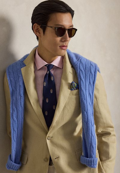 Homme portant des lunettes de soleil, blazer beige avec un mouchoir de poche bleu à motifs, chemise rose, cravate bleu marine et pull bleu clair drapé sur les épaules.