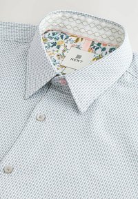 Hellblaues, gemustertes Hemd mit Kragen, das mit einem blumigen Innenfutter, pinkfarbenen Knopfakzenten und einem markierten Etikett "NEXT" versehen ist.