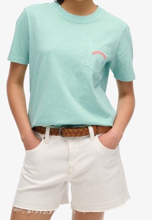 Person iført en lyseblå kortærmet t-shirt med brystlomme og teksten "SUPERDRY", hvide denimshorts og et brunt flettet bælte.