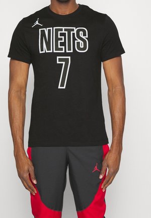 Mężczyzna w czarnej koszulce z tekstem "NETS 7" oraz czarno-czerwonych spodniach z logo Jordan na udzie, stojący na tle jednolitego tła.