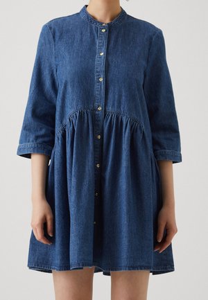 Kvinde iført en blå denimkjole med knapper, trekvartærmer og en indsnævret talje, stående mod en enkel lys baggrund.