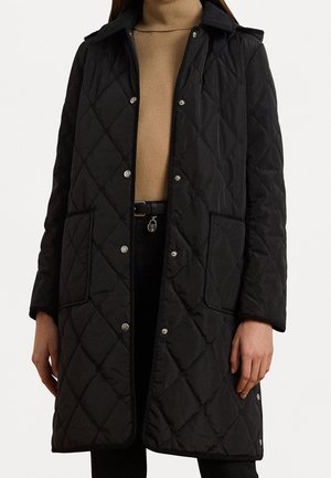 Manteau noir matelassé avec grandes poches, boutons-pression argentés, porté sur un col roulé camel et une ceinture noire avec un pendentif rond.