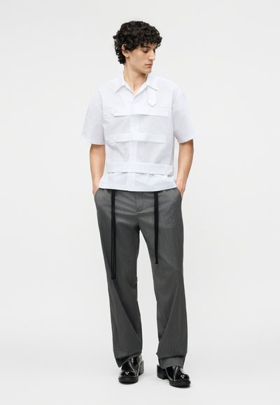 Άνδρας με σγουρά μαλλιά που φοράει άσπρο κοντομάνικο πουκάμισο με λουριά μπροστά, γκρι παντελόνι με ριγέ λεπτομέρειες και μαύρους μακριούς δεσμούς, και μαύρα παπούτσια, στέκεται με τα χέρια στις τσέπες.