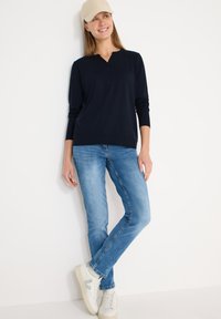 Haut à manches longues bleu marine avec un col en V, associé à un jean bleu clair et des baskets blanches. Le mannequin porte également une casquette beige, debout contre un mur blanc.