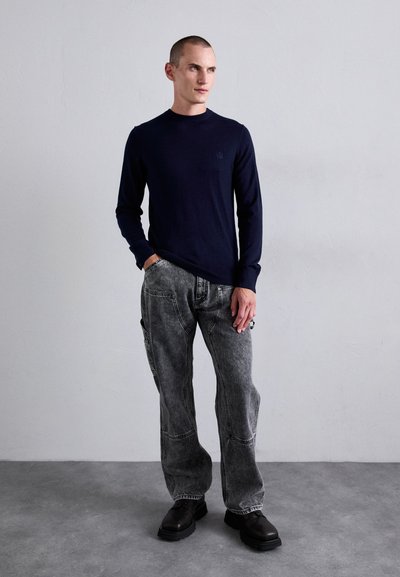 Man wearing a navy long-sleeve sweater, gray acid-wash jeans, and black shoes stands against a plain light gray wall.