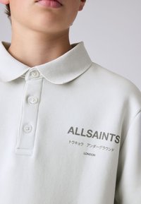 Mladá osoba nosící bílé polo tričko s nápisem "ALLSAINTS" a japonským textem nad nápisem "LONDON" na hrudi.