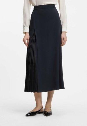 Kobieta w czarnej spódnicy midi z plisowanym detalem po jednej stronie, białej bluzce z długim rękawem i czarnych spiczastych butach typu slingback.