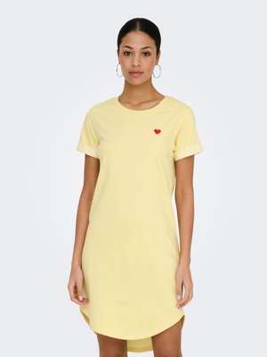 Woman wearing a short-sleeve yellow dress with a small red heart on the chest, standing against a plain light background.