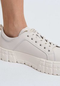 Lichtbeige sneaker met een bovenwerk van synthetisch leer, dikke structuurzool, ronde neus en platte veters. Kenmerkt zich door stiksels en veterogen.