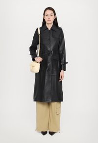 Svart skinn trenchcoat med belte i midjen, store frontlommer og knappelukking. Kombinert med beige cargo-bukser og en liten kremfarget håndveske.
