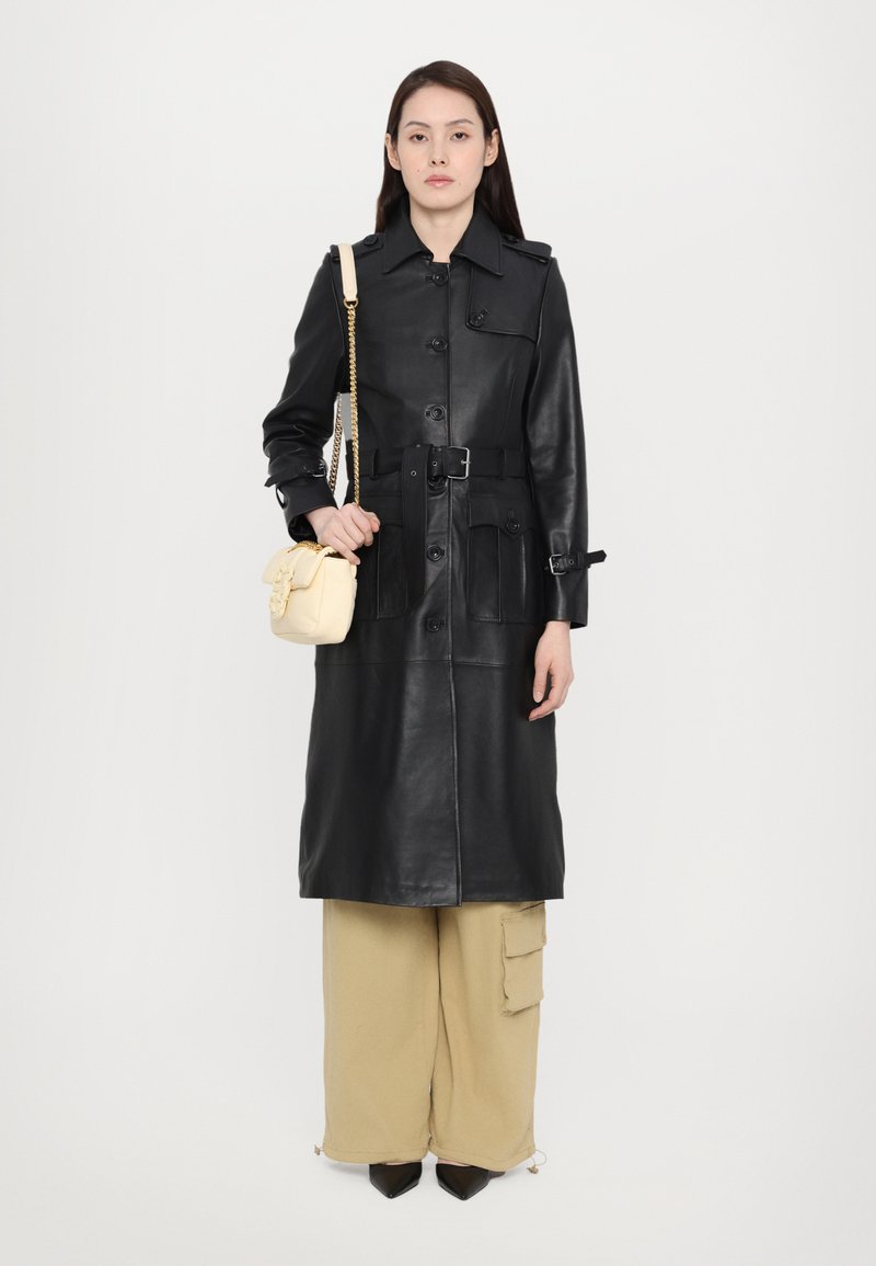 Svart skinn trenchcoat med belte i midjen, store frontlommer og knappelukking. Kombinert med beige cargo-bukser og en liten kremfarget håndveske.