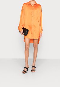 Vestido de satén naranja con botones y dobladillo asimétrico, combinado con tacones negros de tiras y un clutch negro texturizado.