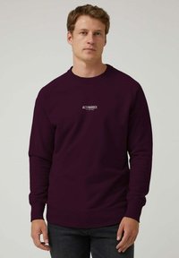 Hombre de pie con un sudadera oscura de color púrpura que tiene el texto "ALTONADOCK es tu camino" en el pecho y jeans oscuros, con un fondo liso.