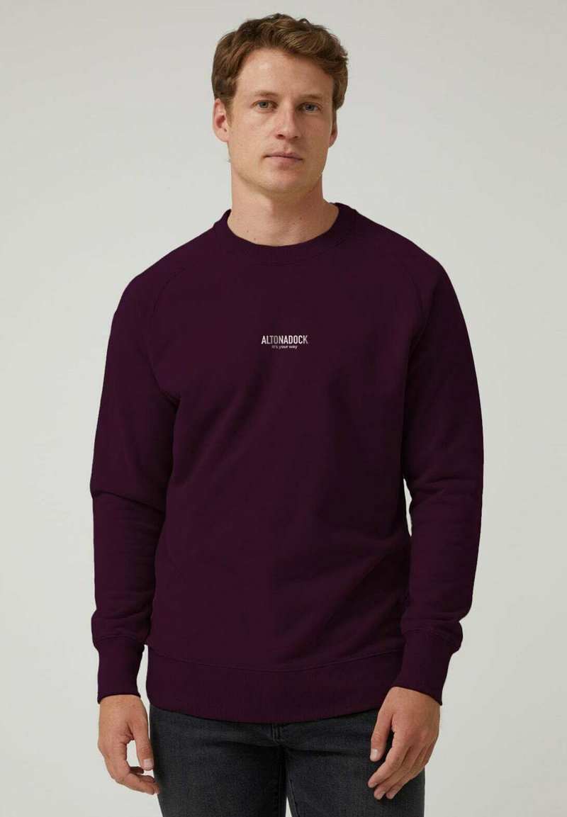 Hombre de pie con un sudadera oscura de color púrpura que tiene el texto "ALTONADOCK es tu camino" en el pecho y jeans oscuros, con un fondo liso.