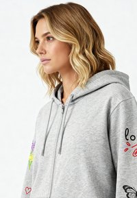 Szürke zipzáras kapucnis pulóver, mely színes hímzéseket tartalmaz virágokról, egy pillangóról és díszes feliratokról az ujjakon.