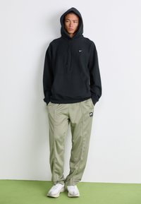 Ung mann iført svart Nike hettegenser, beige Nike joggebukser og hvite joggesko som står mot en hvit vegg på grønn gulv.